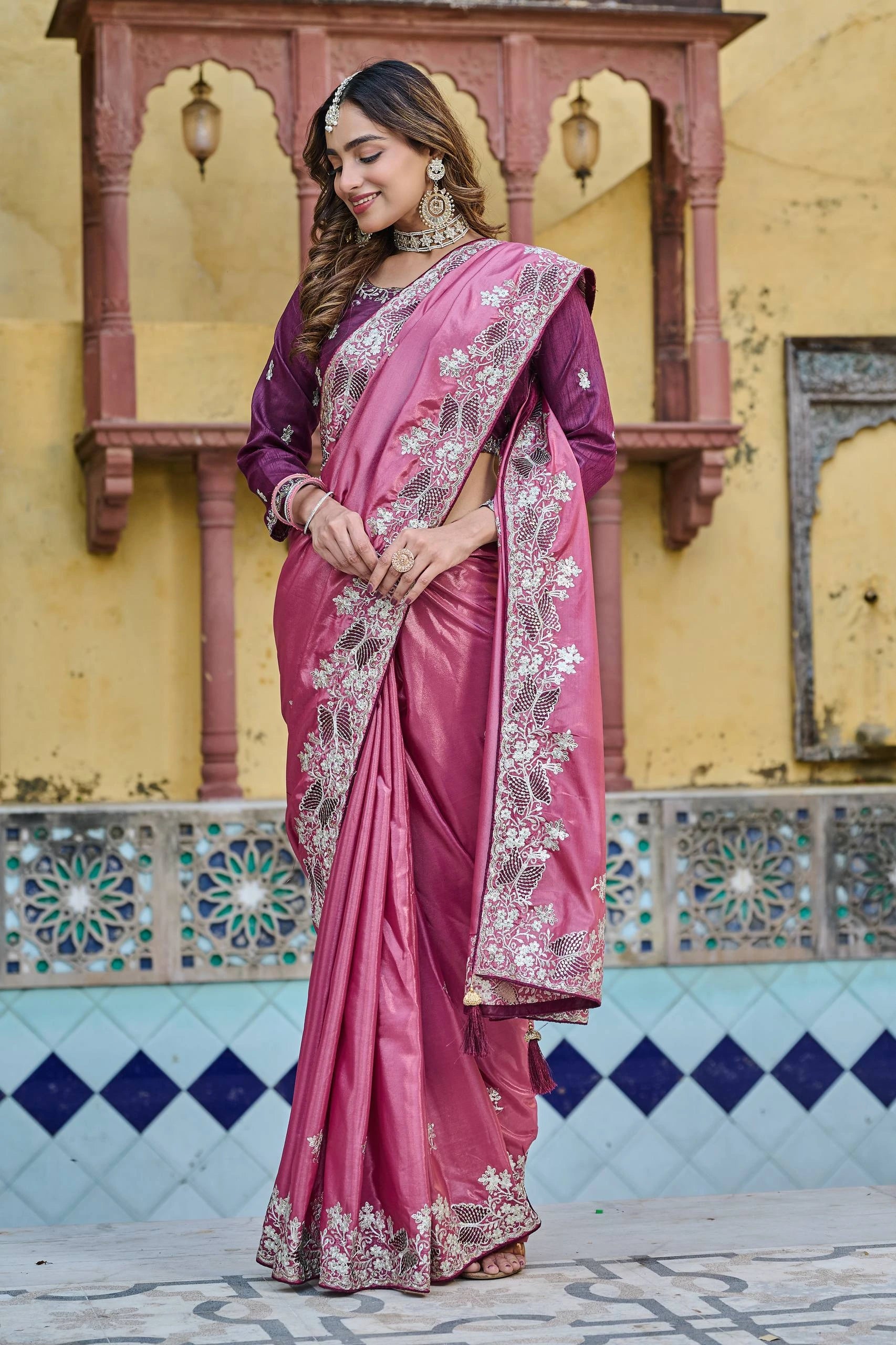 Woman in a pink saree with intricate patterns standing in front of a decorative wall.